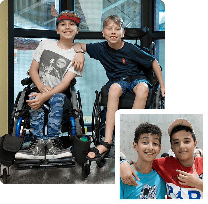 Two boys in a wheelchair and one boy is holding his hand up to the side of their head.