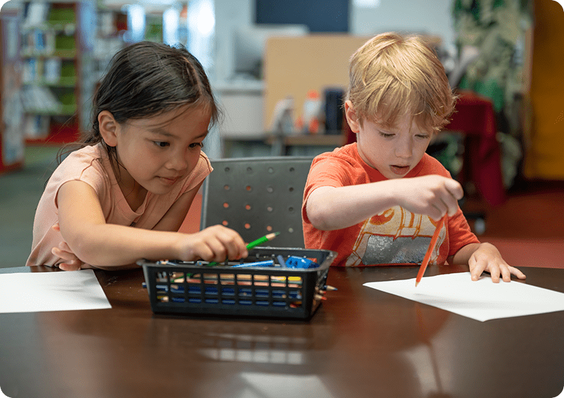 Two children are sitting at a table and one is drawing.